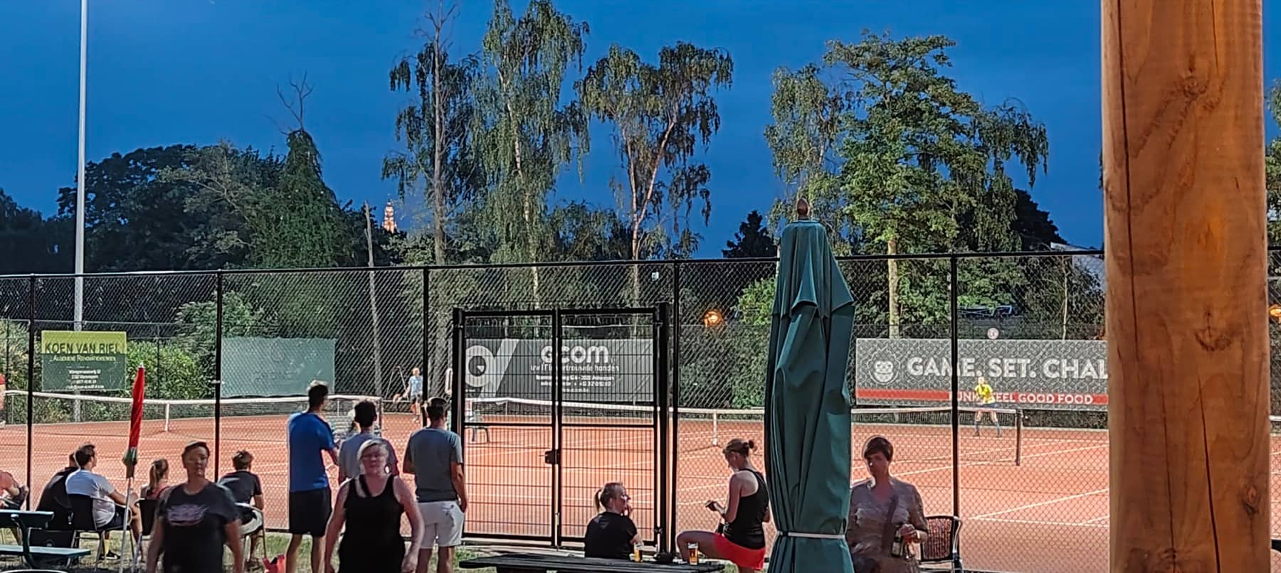Leden aan het terras bij valavond, met achter het hek spelers op de verlichte gravelterreinen