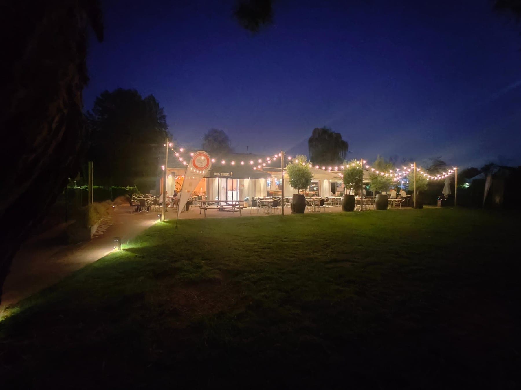 Het clubhuis bij nacht, met lichtslingers boven het terras en het gazon ervoor