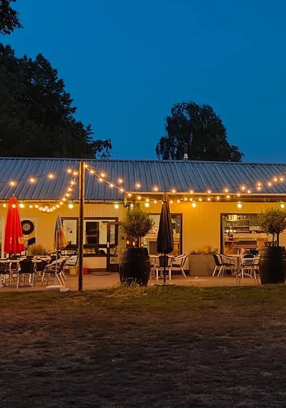 Het clubhuis in het blauwe uur, met lichtslingers boven het terras en verlichte terreinen ernaast