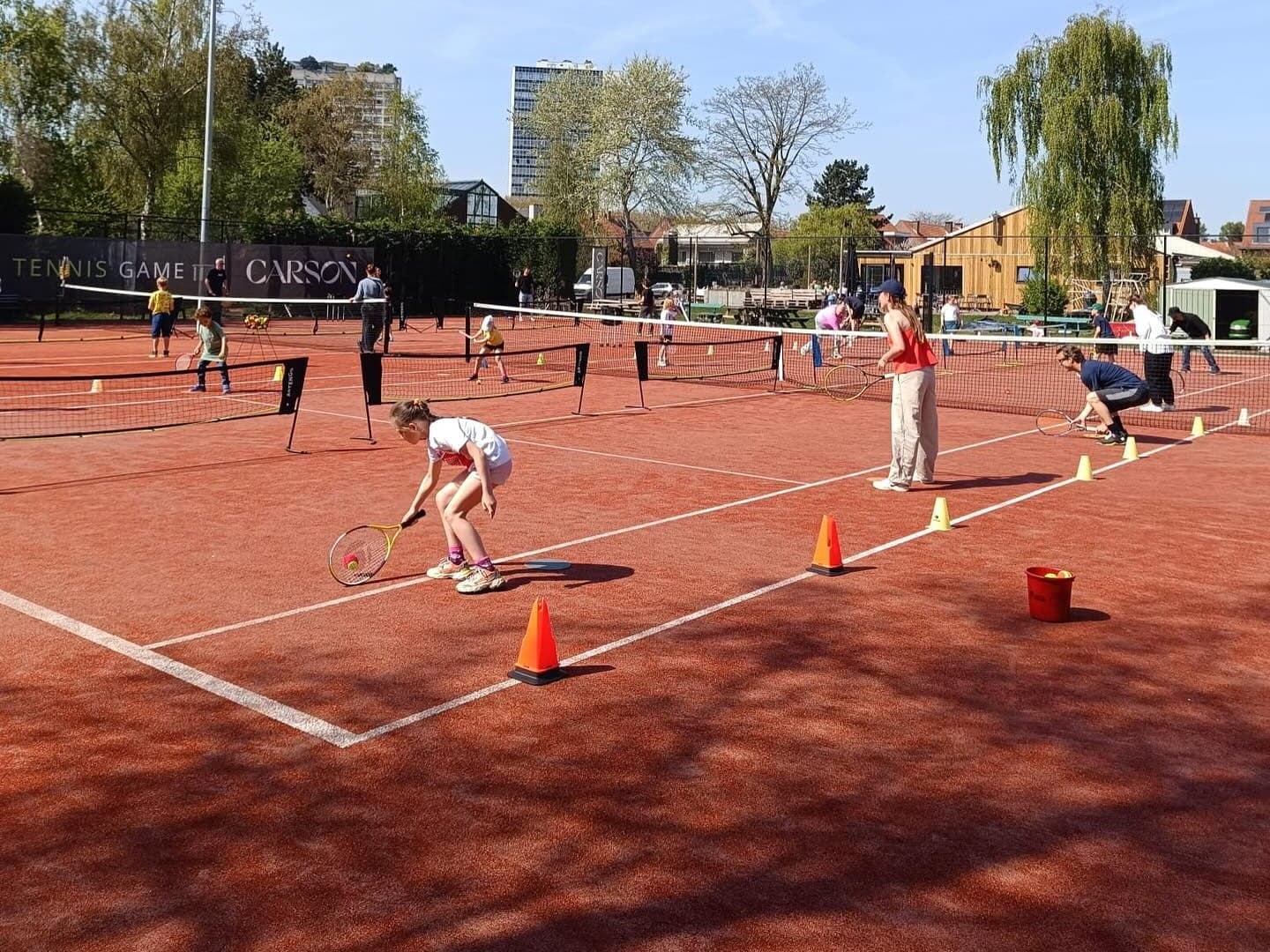 Kinderen in de tennisles op het gravel, met lage netten, pylonen en een ballenemmer