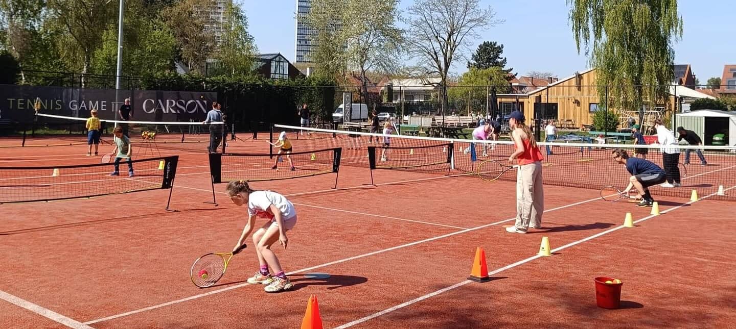 Kinderen in de tennisles op het gravel, met lage netten, pylonen en een ballenemmer