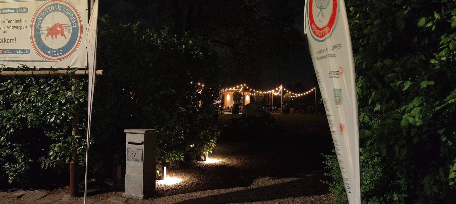 De ingang van de club bij avond, met het clubbord en verlichte paden naar het terras